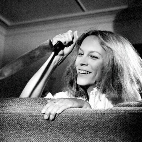 An image of Jamie Lee Curtis as Laurie Strode in the movie Halloween (1978). She is smiling as she peaks over the back of a couch with a large kitchen knife in hand.